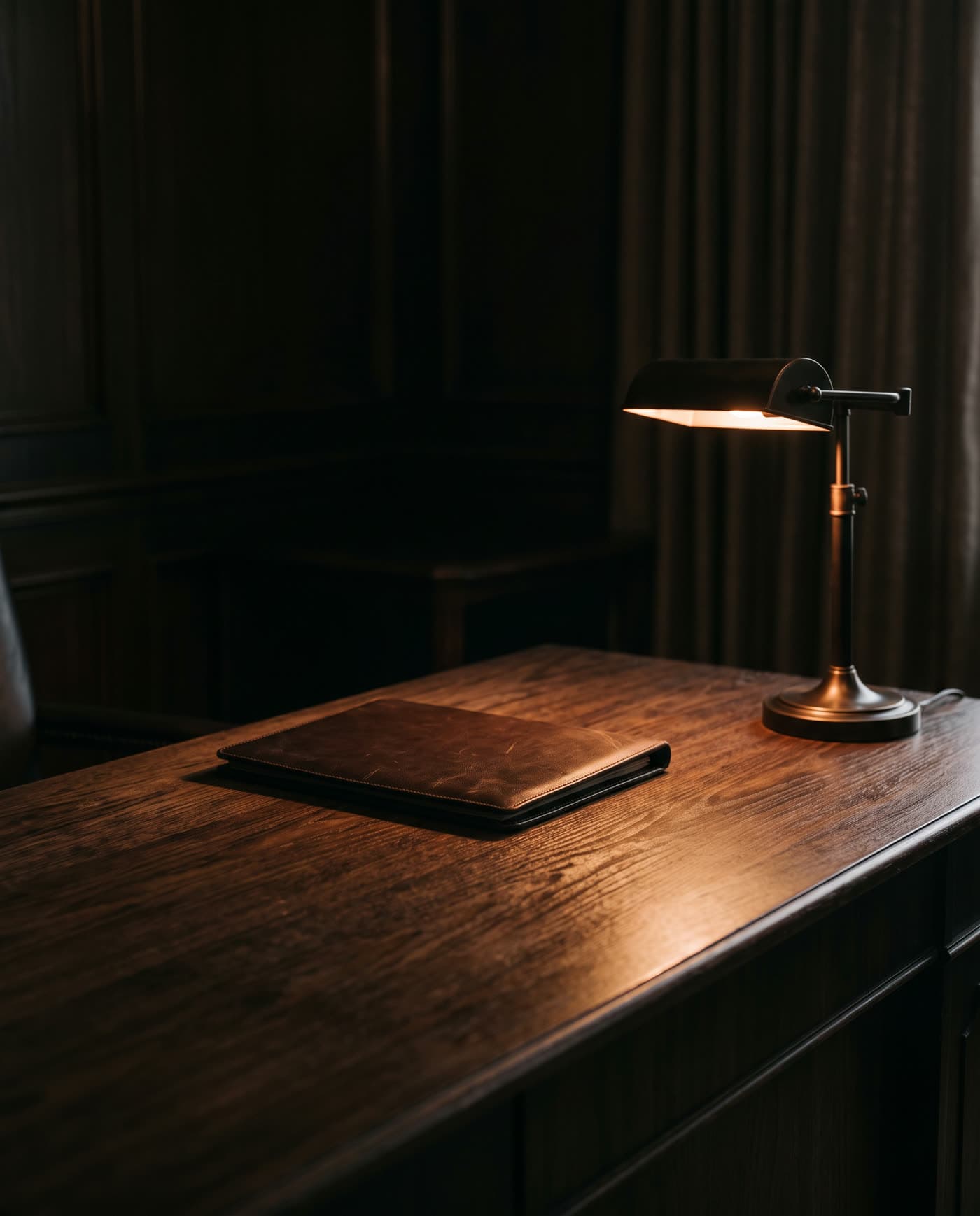 Escritorio de madera oscura con una carpeta de cuero cerrada, iluminado por una lámpara de bronce en una oficina en penumbra.